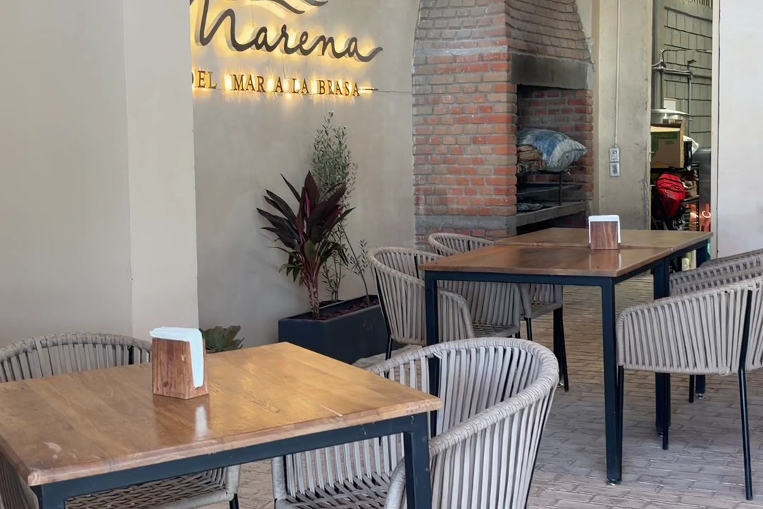 Salón interior con mesas de madera, sillas de rattan y el muro de ladrillo del horno.