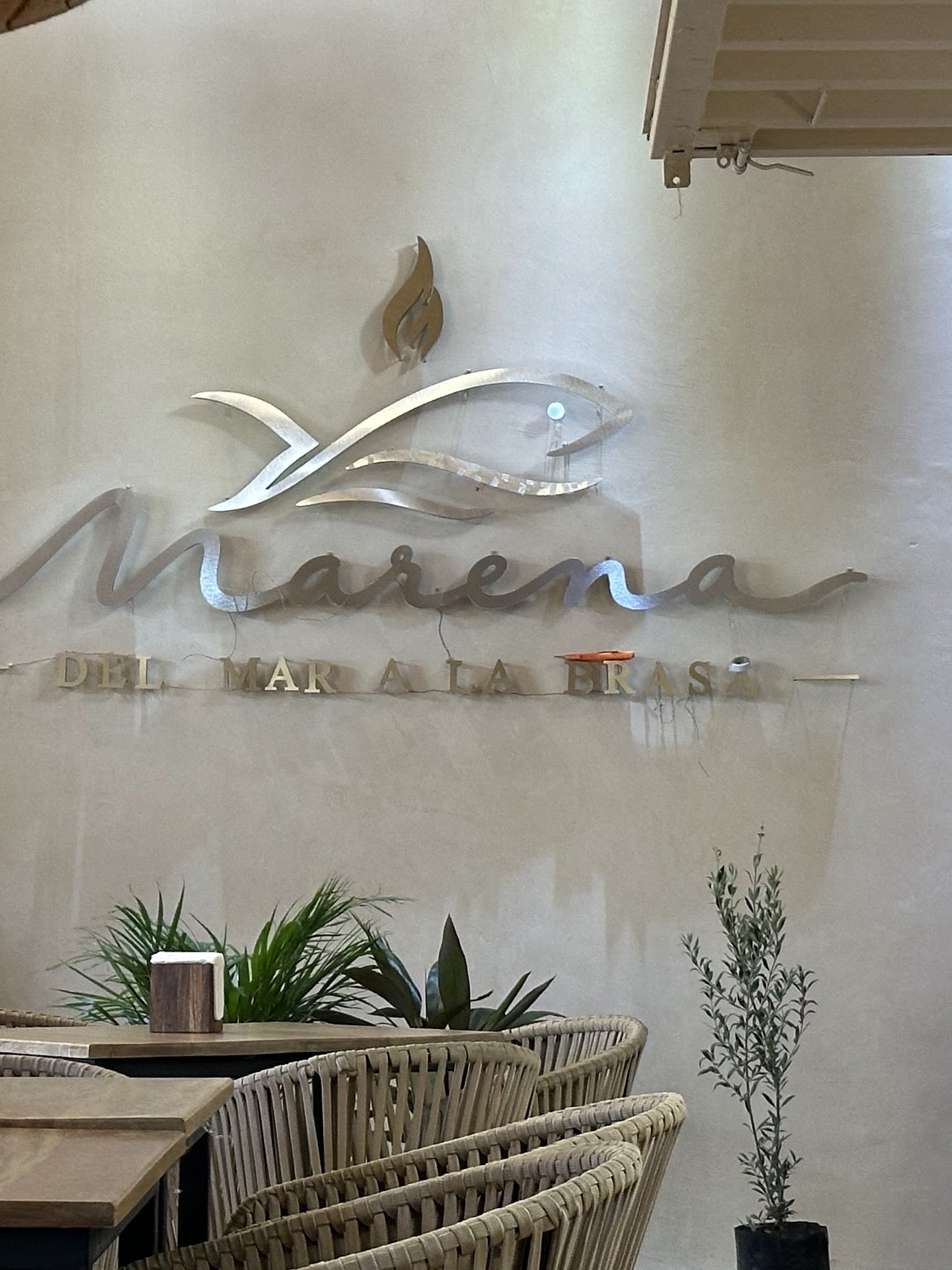 El logotipo de Marena en relieve sobre el muro, con plantas colgantes y las mesas del salón al fondo.