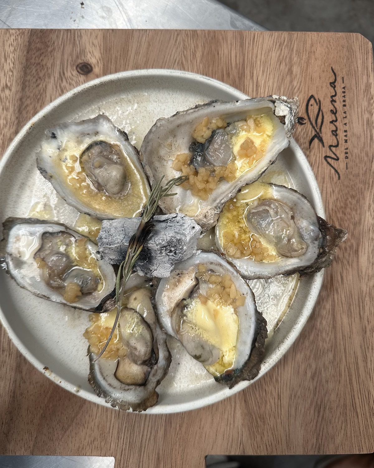 Ostiones frescos abiertos y gratinados, servidos con romero sobre plato de la casa.